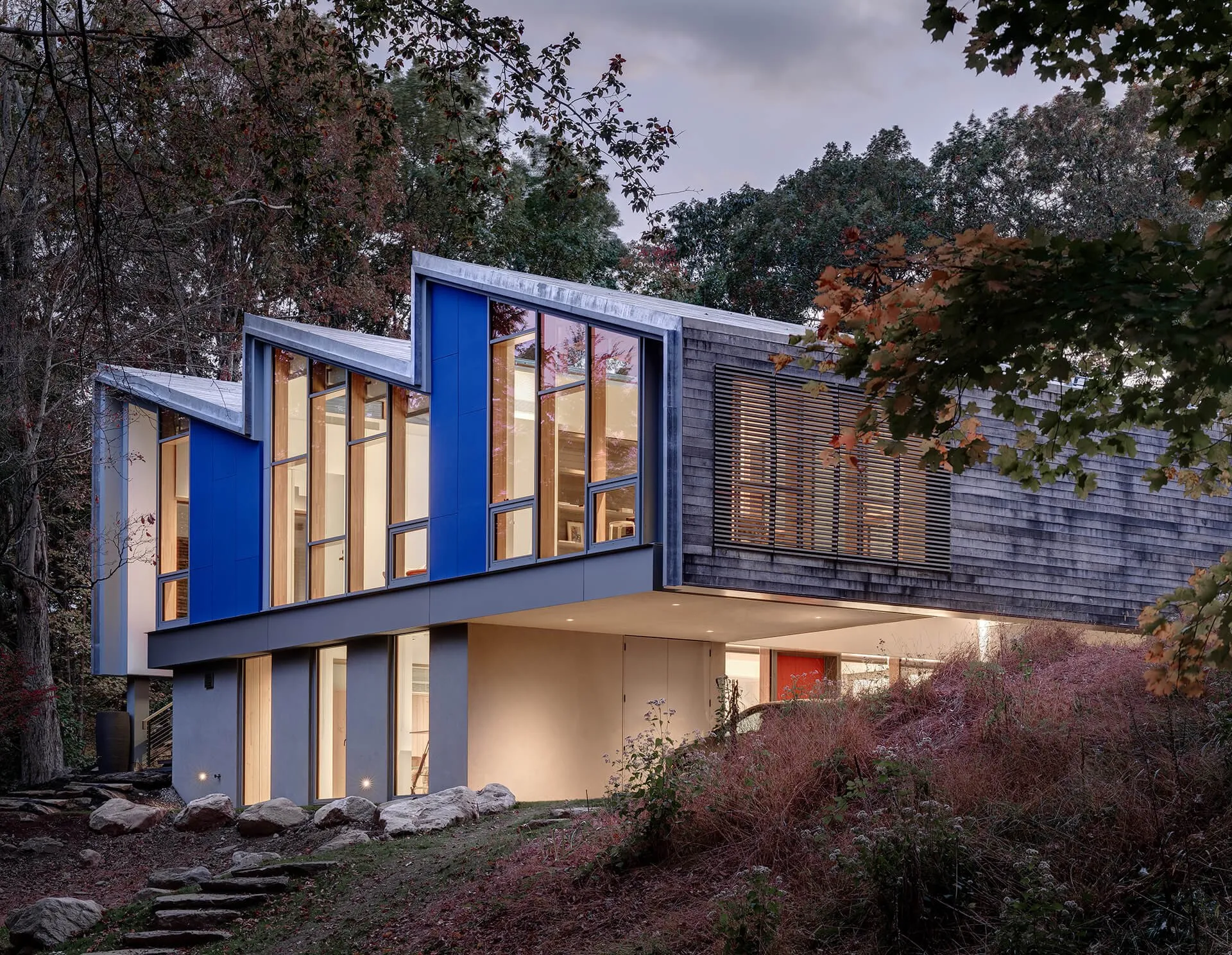 Modern house with a sawtooth roofline, blue panels, and tall lit windows cantilevered over a wooded hillside at dusk — cover image for the e2 Engineers website project