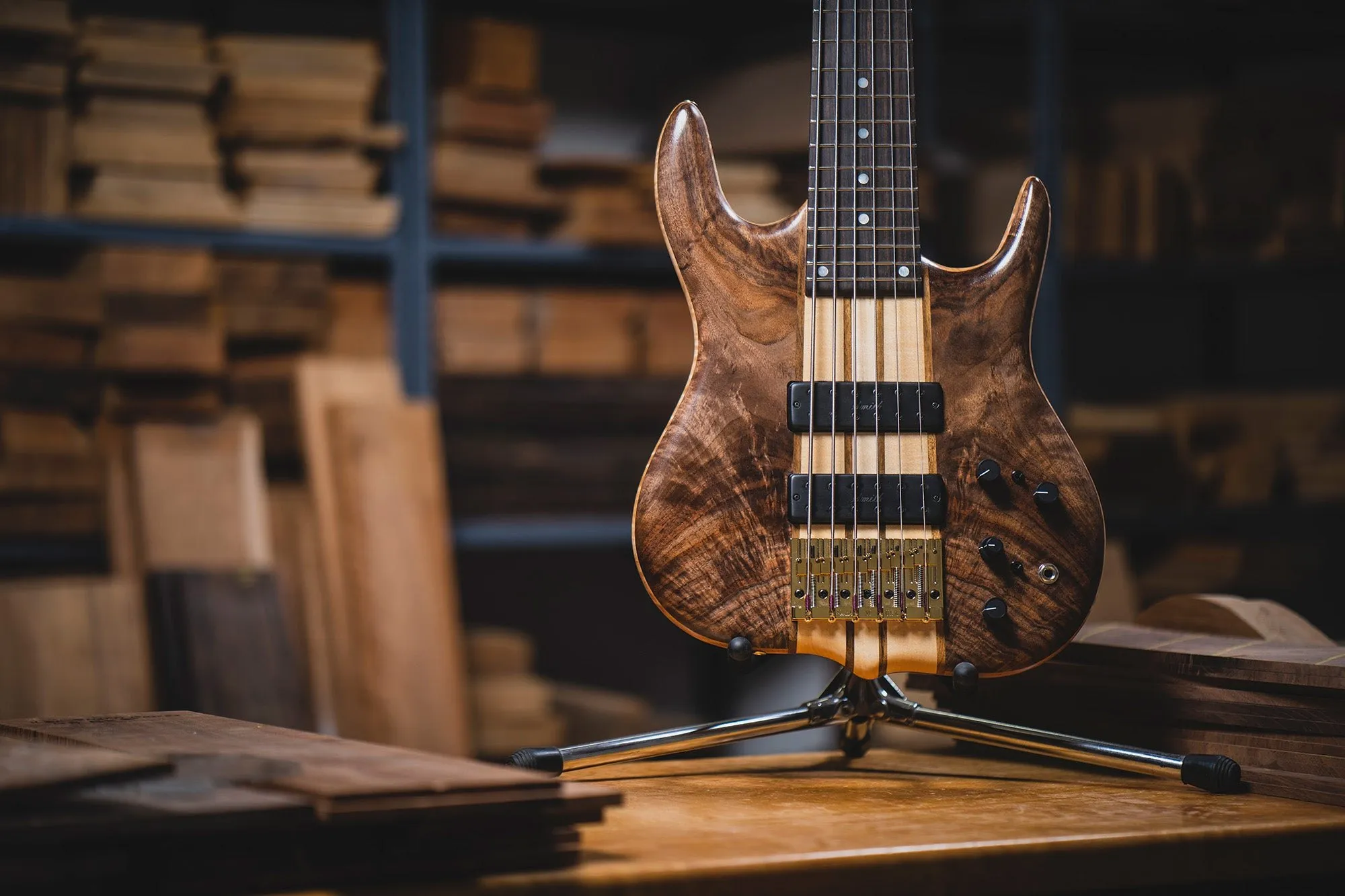 Handmade multi-string bass with a figured burl body on a stand in a workshop stacked with tonewood — cover image for the Ken Smith Basses website project